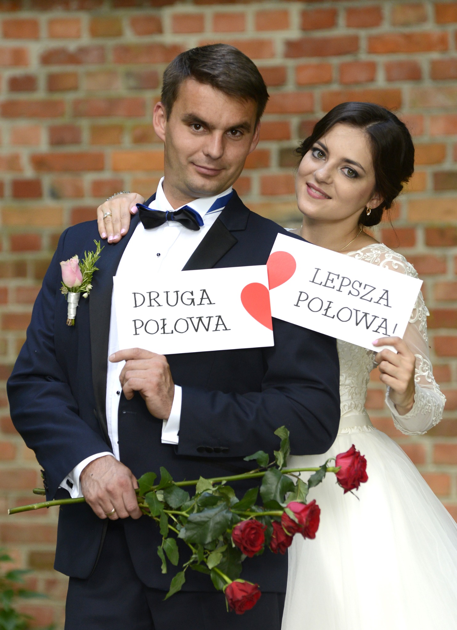 Uśmiechnięta para młoda trzymająca kartki z napisem 'Druga Połowa' i 'Lepsza Połowa!' oraz czerwone róże na tle ceglanej ściany.