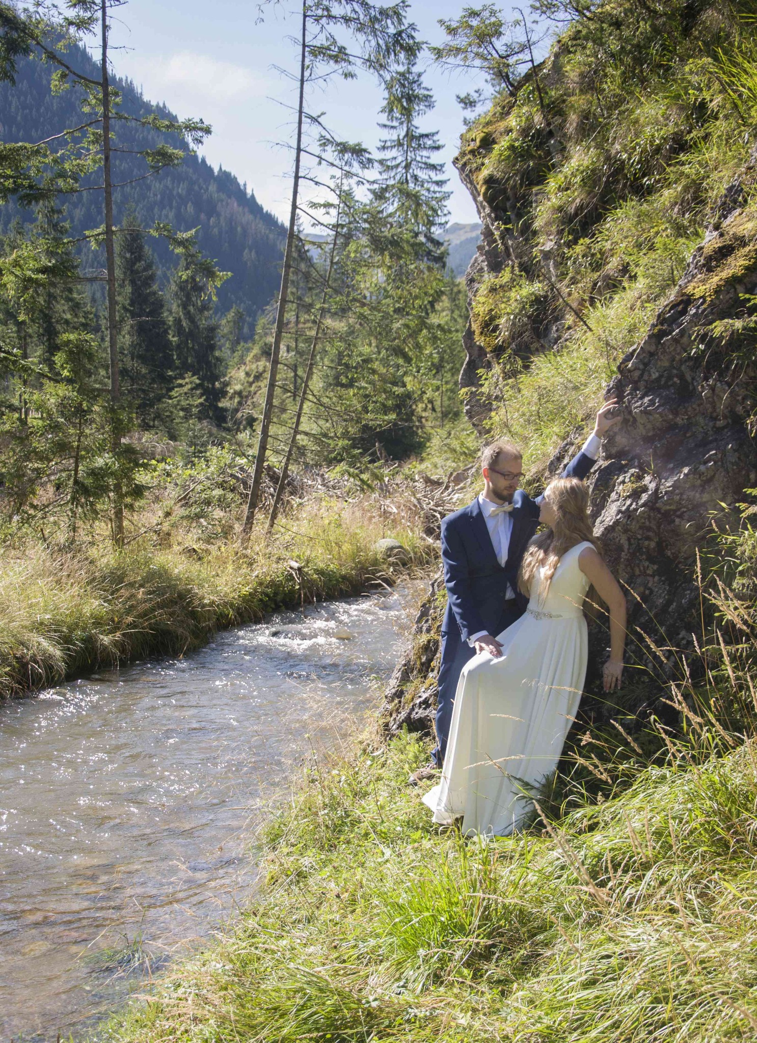 Para młoda w plenerze górskim: panna młoda w białej sukni opiera się o skałę, pan młody w granatowym garniturze, obok płynie strumień, w tle las i góry.