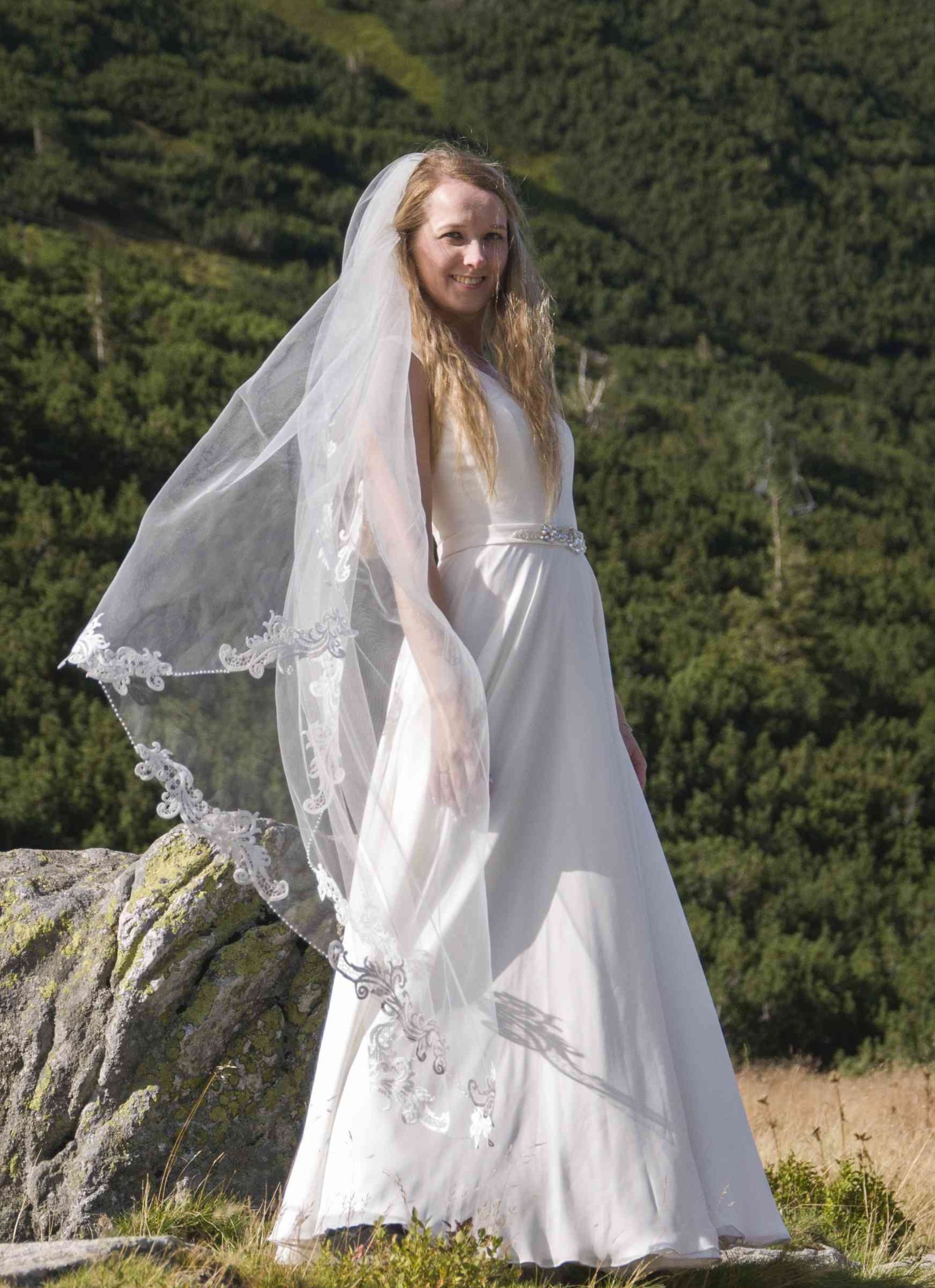 Panna młoda w zwiewnej sukni i welonie z koronkowym brzegiem pozuje na tle górskiego krajobrazu. Uśmiechnięta blondynka na skale.
