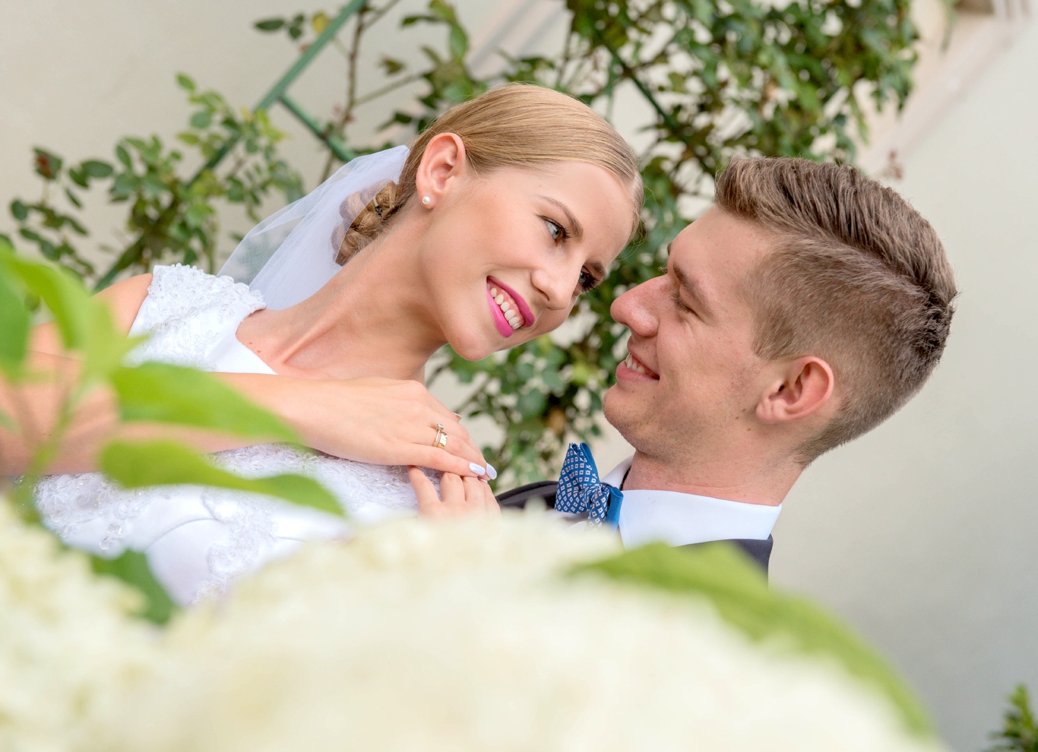 Uśmiechnięta panna młoda w białej sukni i welonie, z delikatnym makijażem, spogląda na pana młodego w muszce na tle zieleni.