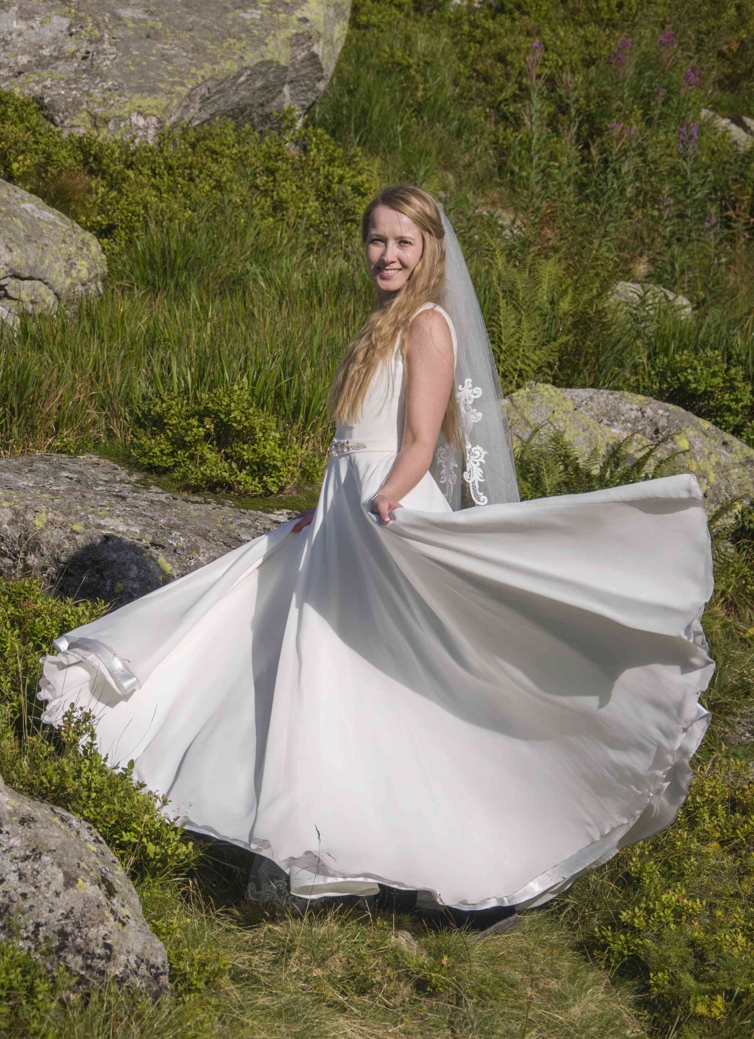 Panna młoda w zwiewnej sukni wiruje na tle górskiej łąki w Łukowie. Delikatny welon i uśmiech dodają uroku. Kamienie i zieleń w tle podkreślają naturalny charakter.