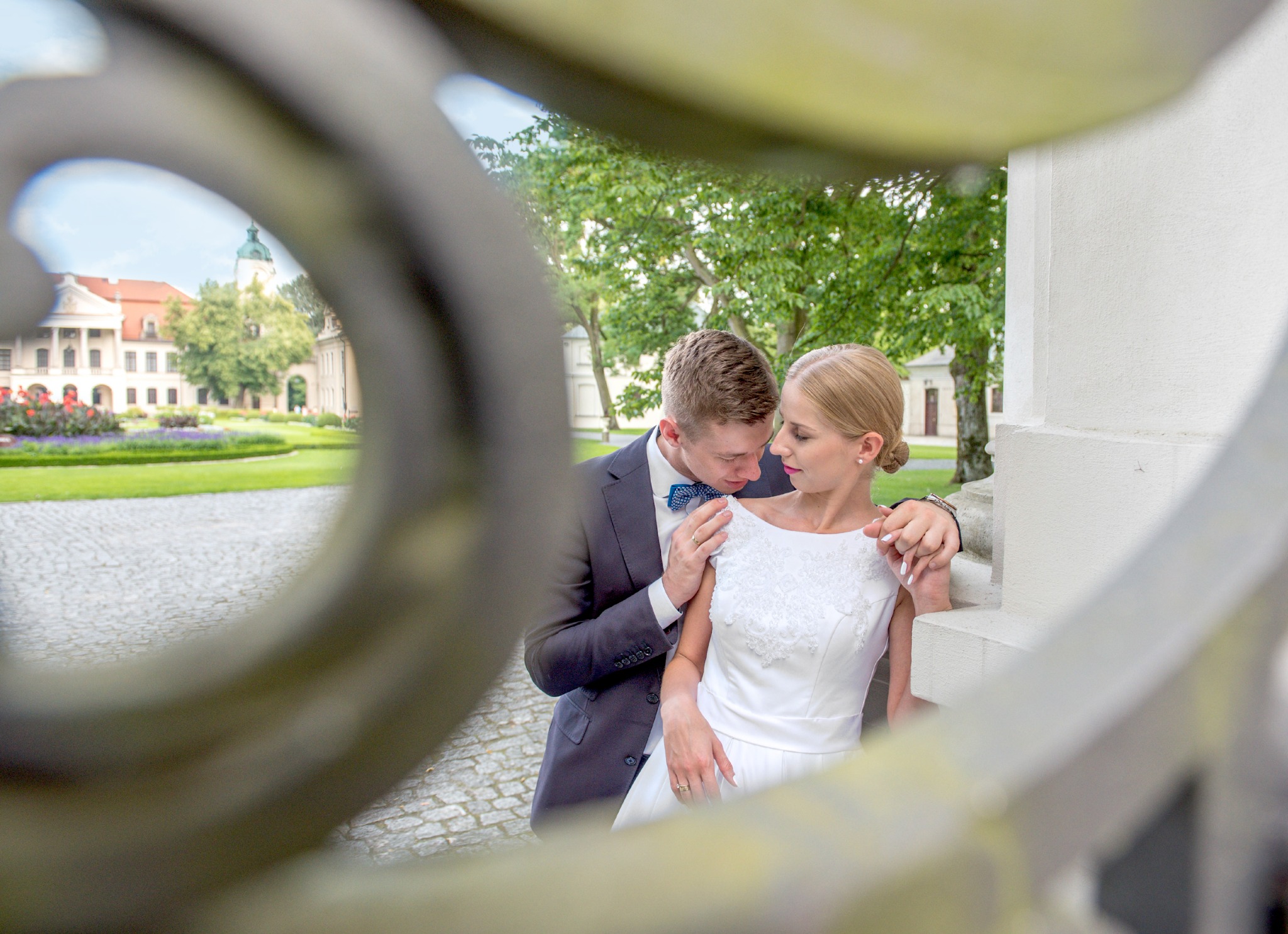 Elegancka para młoda w objęciach, uchwycona przez ozdobne ogrodzenie, z pałacem w tle. Pan młody w ciemnym garniturze i muszce, panna młoda w białej sukni.