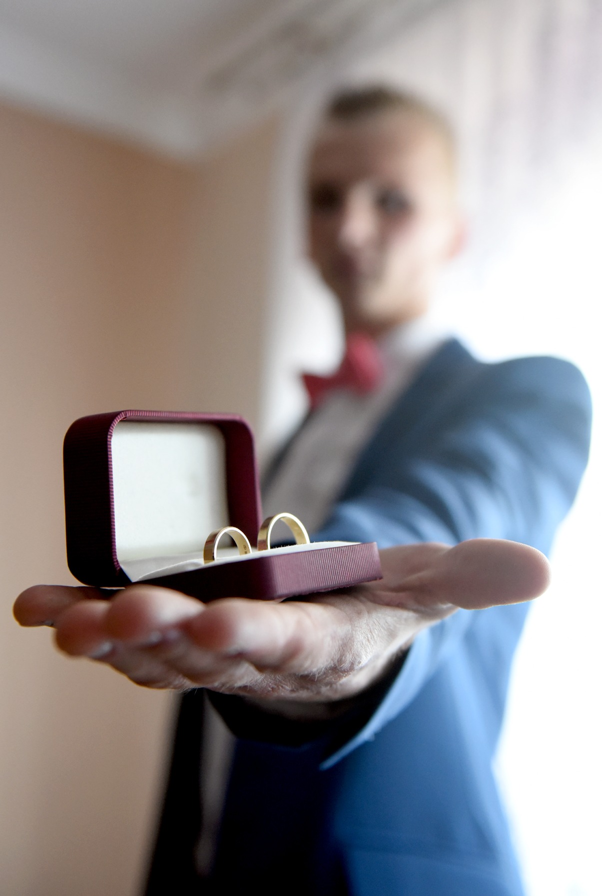 Mężczyzna w błękitnej marynarce prezentuje otwarte pudełko z złotymi obrączkami na dłoni. Tło rozmyte, widoczna biała koszula i czerwona muszka.