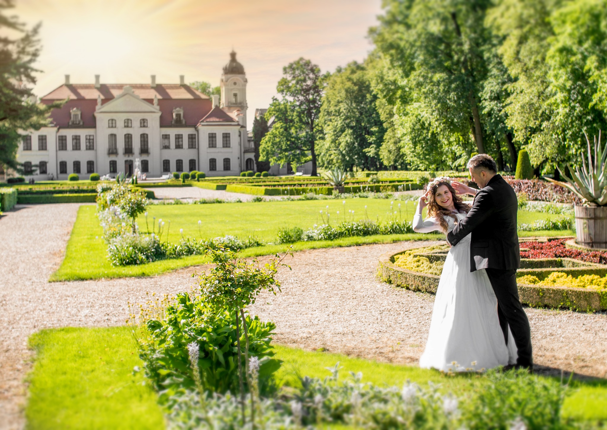 Para młoda w objęciach na tle pałacu i zieleni. Panna młoda w białej sukni i wianku, pan młody w czarnym garniturze. Słoneczny dzień, parkowe otoczenie.