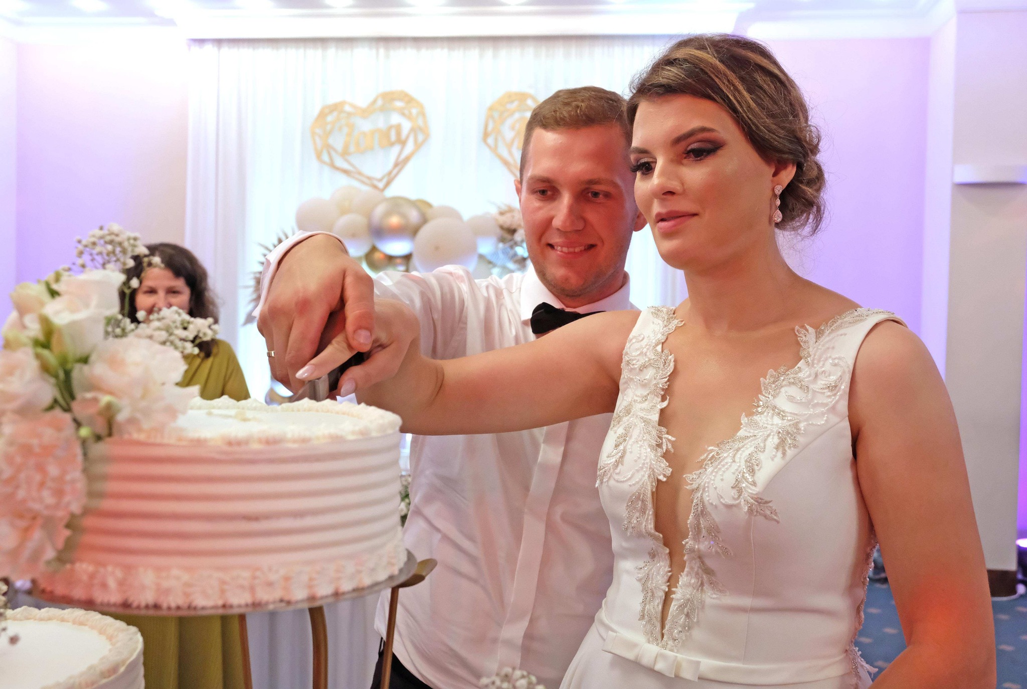 Uśmiechnięta para młoda kroi tort weselny, ozdobiony kwiatami, w jasnym wnętrzu sali bankietowej z dekoracjami.