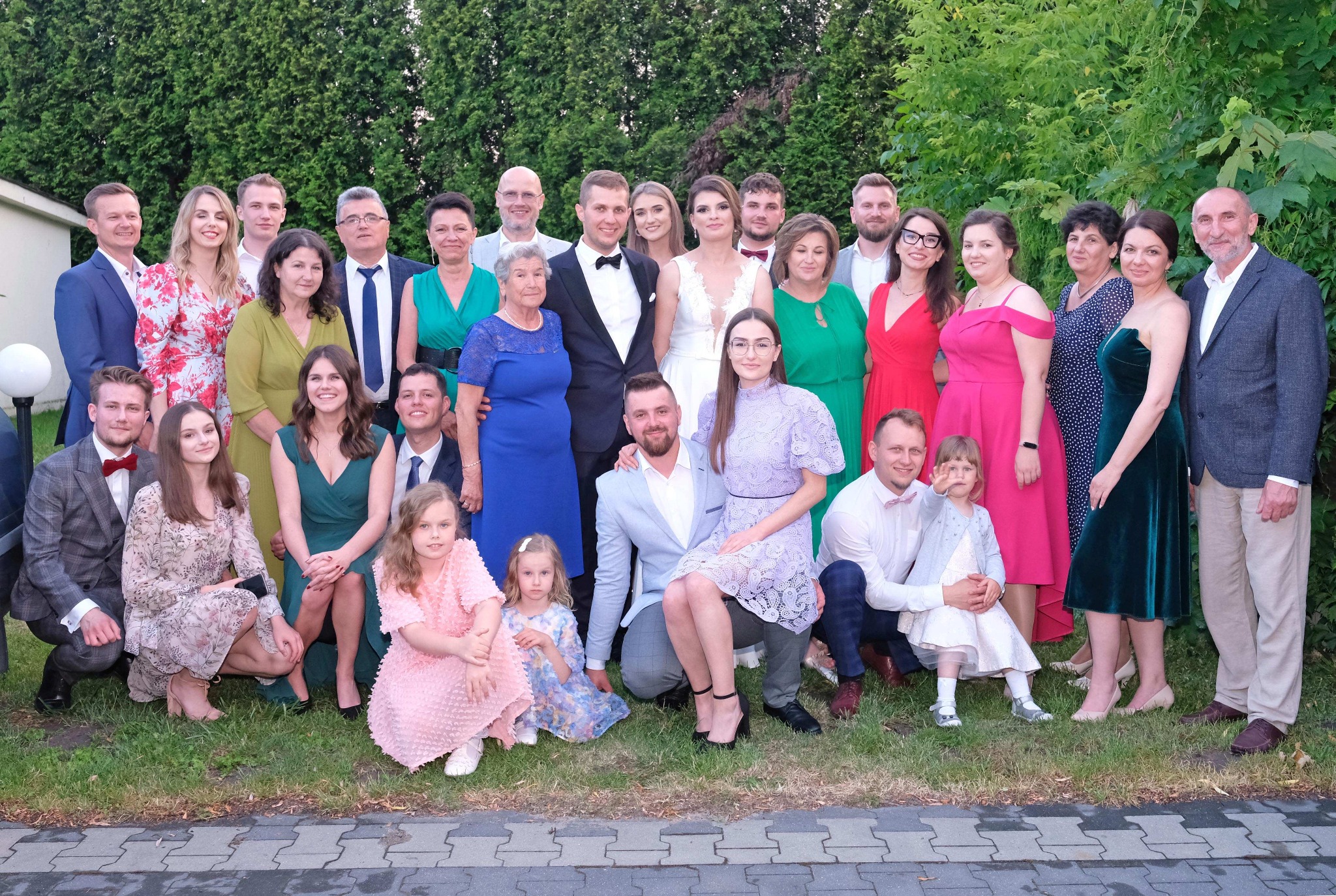 Grupowe zdjęcie ślubne z panną młodą i panem młodym w centrum, otoczonych przez gości w różnorodnych strojach na tle zieleni.