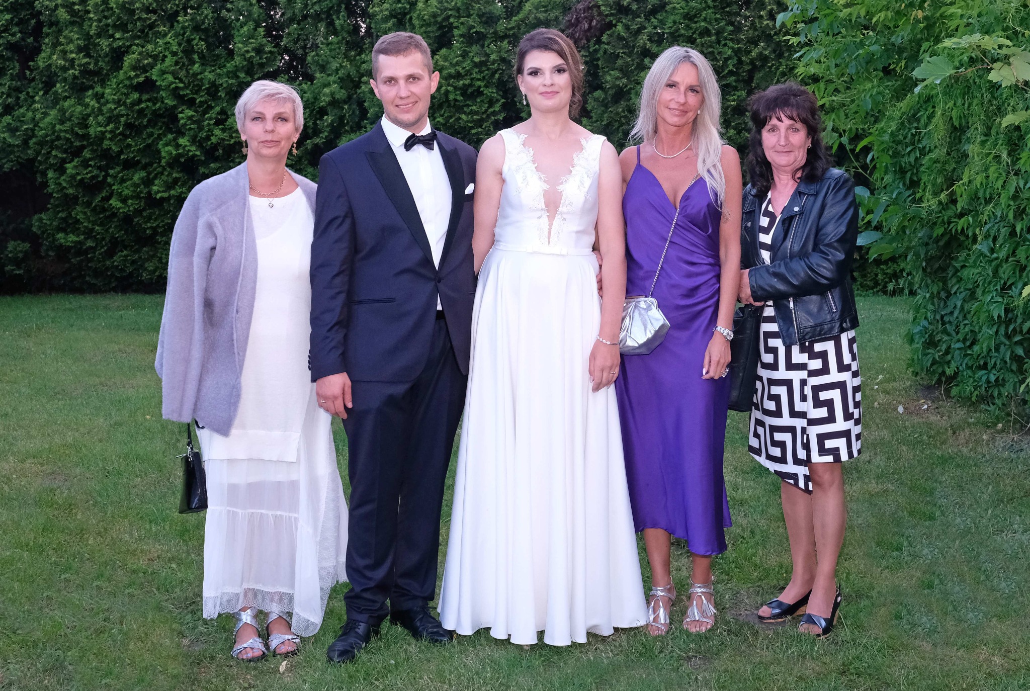 Portret grupowy: para młoda w bieli i garniturze, otoczona trzema kobietami w sukienkach na tle zieleni. Lekkie uśmiechy, naturalne światło.