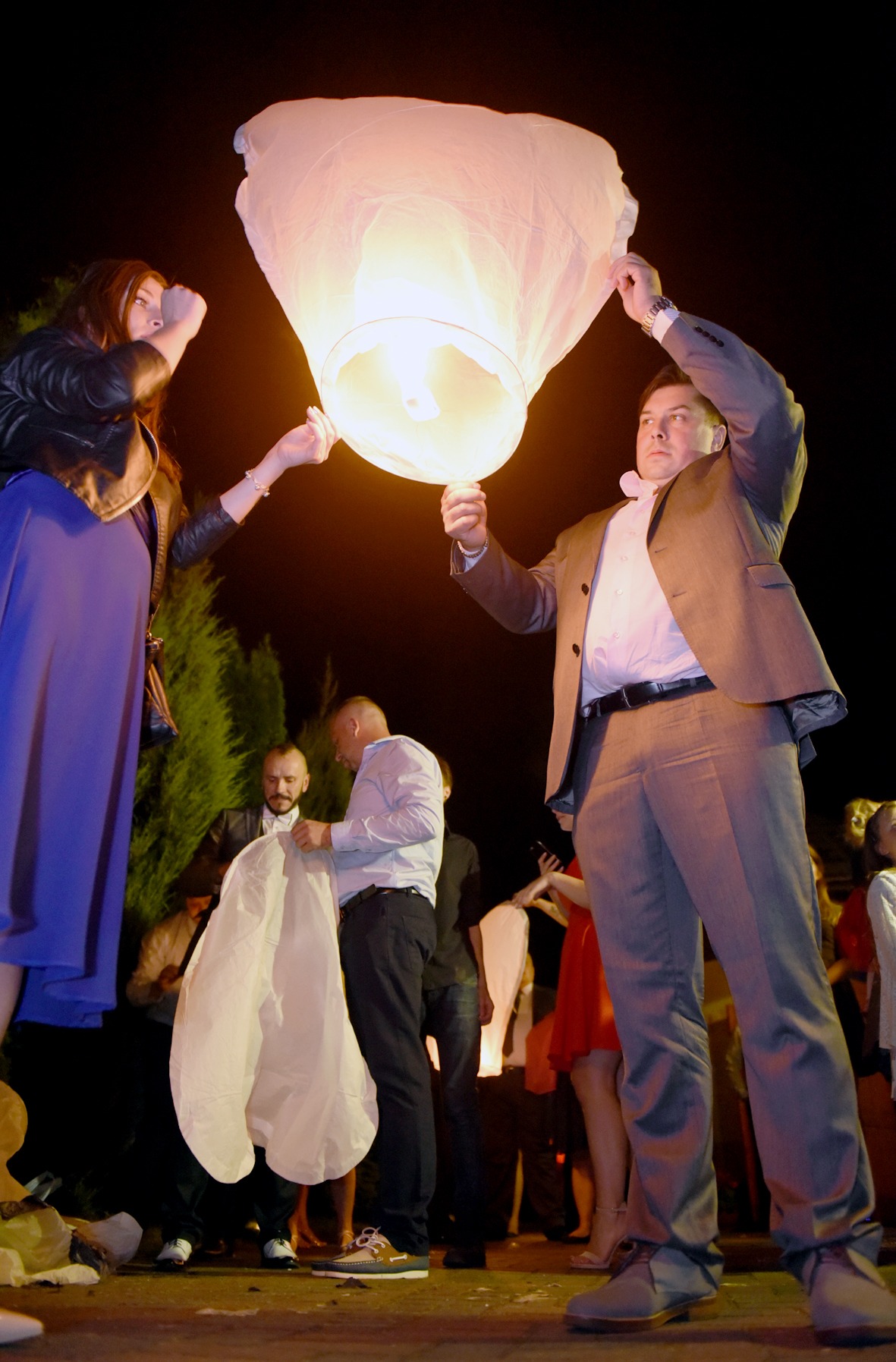Para w strojach wieczorowych wypuszcza lampion szczęścia nocą. Ujęcie z dołu, widoczni goście i fragmenty zieleni w tle, budując kameralny nastrój.