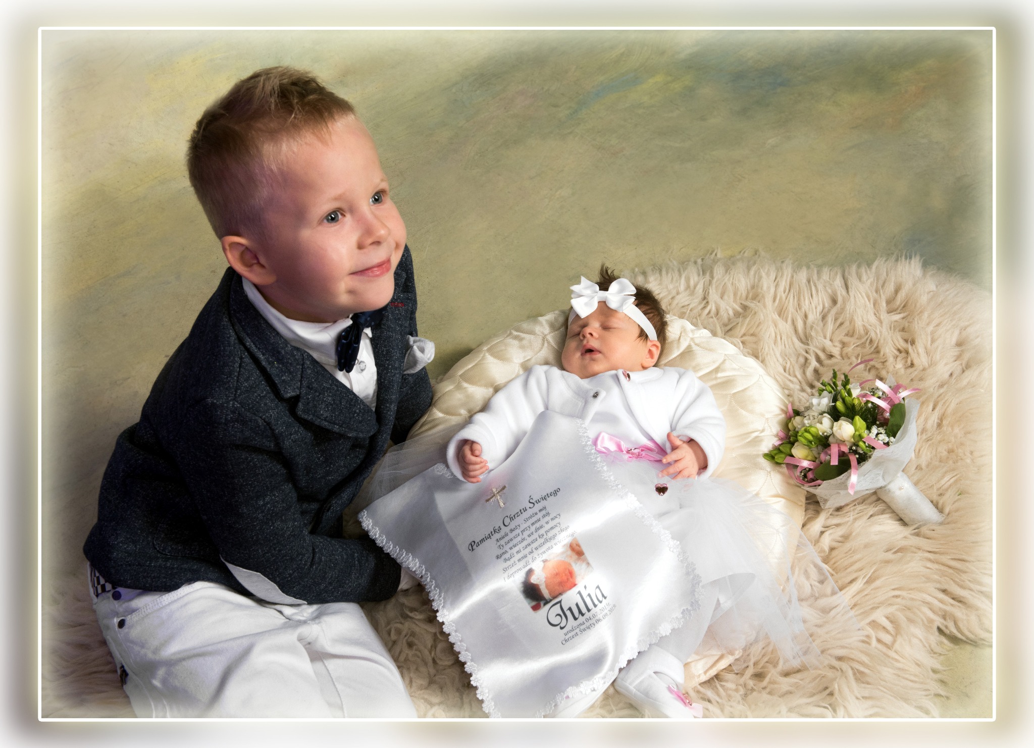 Uśmiechnięty chłopiec w garniturze spogląda na śpiącą dziewczynkę z kokardą i bukiecikiem, leżącą na poduszce z pamiątką chrztu. Pastelowe tło.