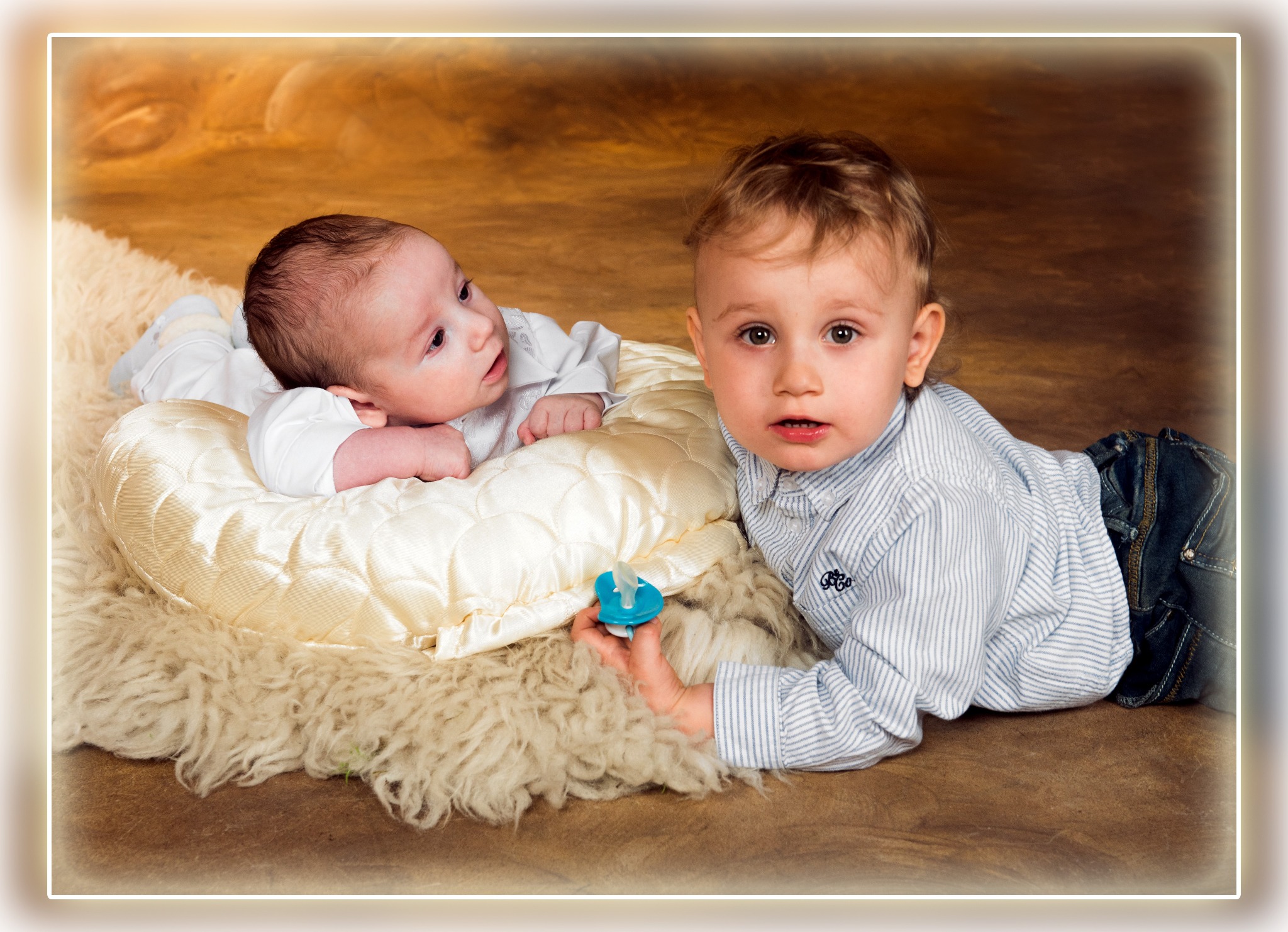 Dwaj chłopcy leżą na miękkim dywanie w studio. Jeden niemowlak na poduszce, drugi trzyma smoczek i patrzy w obiektyw. Ciepłe, studyjne oświetlenie.