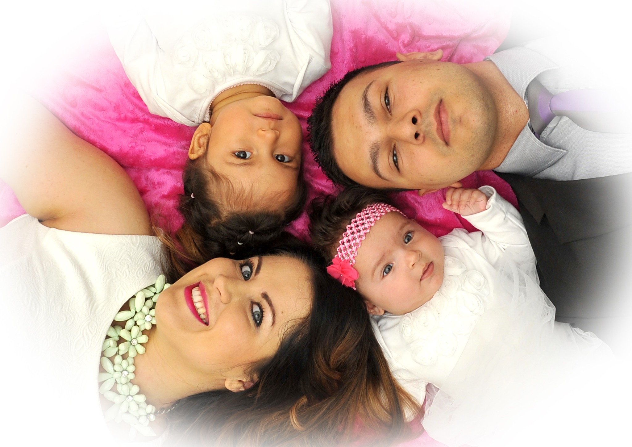 Uśmiechnięta rodzina (mama, tata, dwie córki) leżąca na różowym tle, spojrzenie w górę, ujęcie z góry. Dzieci ubrane na biało, mama w białej sukience.