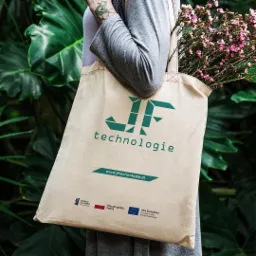 Torba bawełniana z zielonym logo 'JF technologie' i adresem strony internetowej, trzymana przez osobę z tatuażem na tle zielonych liści i kwiat&oacute;w.