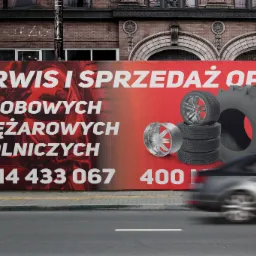 Billboard reklamujący serwis i sprzedaż opon osobowych, ciężarowych i rolniczych z numerem telefonu, na tle przejeżdżający samochód.