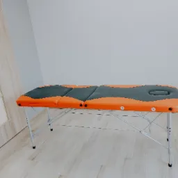 Składany stół do masażu w kolorze pomarańczowo-szarym, z regulowanymi nogami, ustawiony w jasnym pomieszczeniu z drewnianą podłogą i drzwiami w tle.