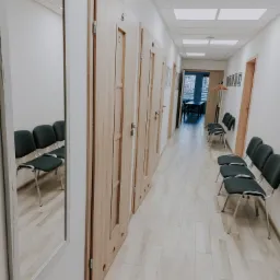Korytarz w placówce medycznej z rzędem drewnianych drzwi do gabinetów, krzesłami dla pacjentów i lustrem.