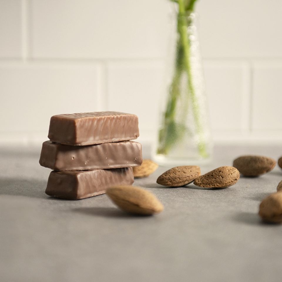 Trzy czekoladowe batony ułożone w stos, otoczone migdałami na szarym tle, z rozmytym wazonem z kwiatami w tle.