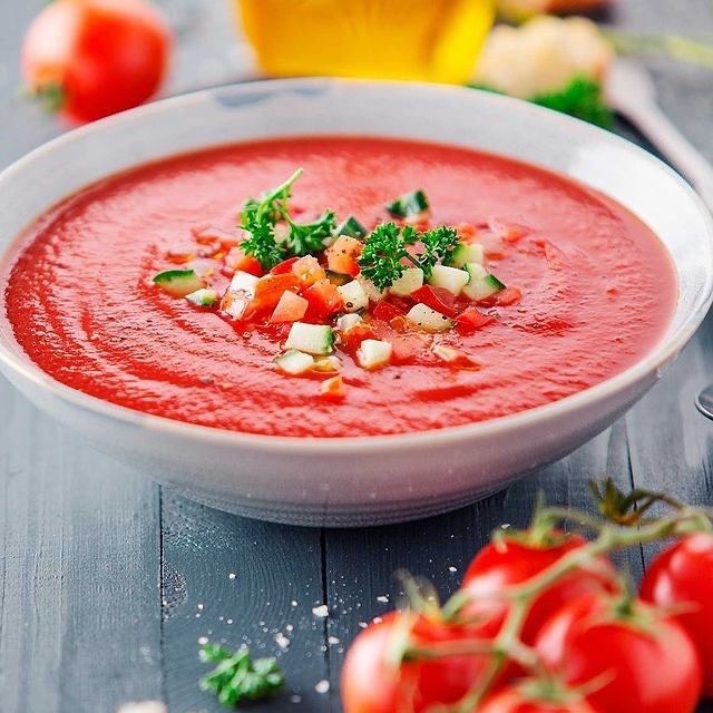 Miseczka z gazpacho, udekorowana posiekanymi pomidorami, ogórkiem i natką pietruszki, obok gałązka świeżych pomidorów na szarym, drewnianym stole.