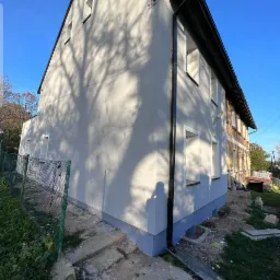 Wykończona elewacja domu dwukondygnacyjnego z widocznym fragmentem starej ceglanej ściany, cień drzewa na świeżo pomalowanej ścianie, rynna spustowa i fragment fundamentu w kolorze niebieskim.