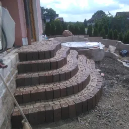 Kaskadowe schody zewnętrzne z kostki brukowej w kolorze ceglastym, prowadzące do tarasu z basenem, widoczne narzędzia budowlane i worki z cementem.