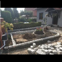 Układanie kostki granitowej wokół rabaty kwiatowej i trawnika przed domem jednorodzinnym, widoczne przygotowane podłoże i zapas kostki brukowej.