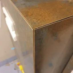 Szczegółowe ujęcie narożnika obudowy z płytek imitujących rdzę, z widocznymi klamrami poziomującymi i fugą w kolorze złota.