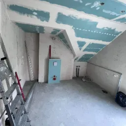 Wnętrze łazienki w trakcie prac wykończeniowych, widoczne płyty kartonowo-gipsowe na suficie i ścianach, instalacja wodna oraz drabina malarska z boku.