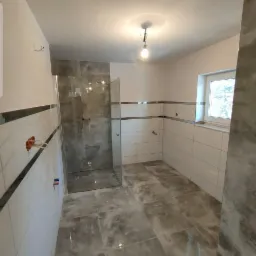 Nowoczesna łazienka z przeszkloną kabiną prysznicową, wykończona płytkami w odcieniach szarości i bieli, z widocznymi wystającymi elementami instalacji wodnej i elektrycznej.