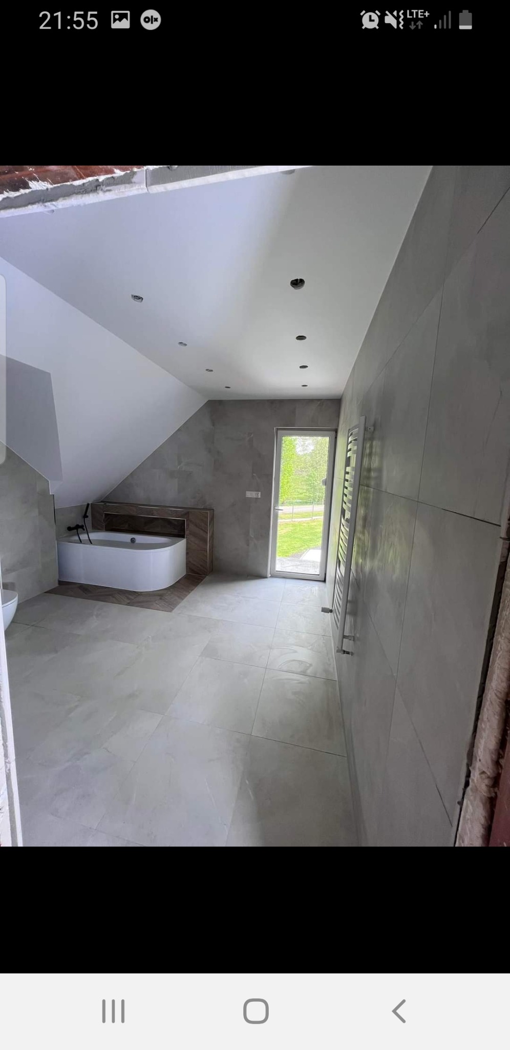 Nowoczesna łazienka na poddaszu z wanną obudowaną drewnem, szarymi płytkami na ścianach i podłodze oraz grzejnikiem drabinkowym obok przeszklonych drzwi balkonowych.