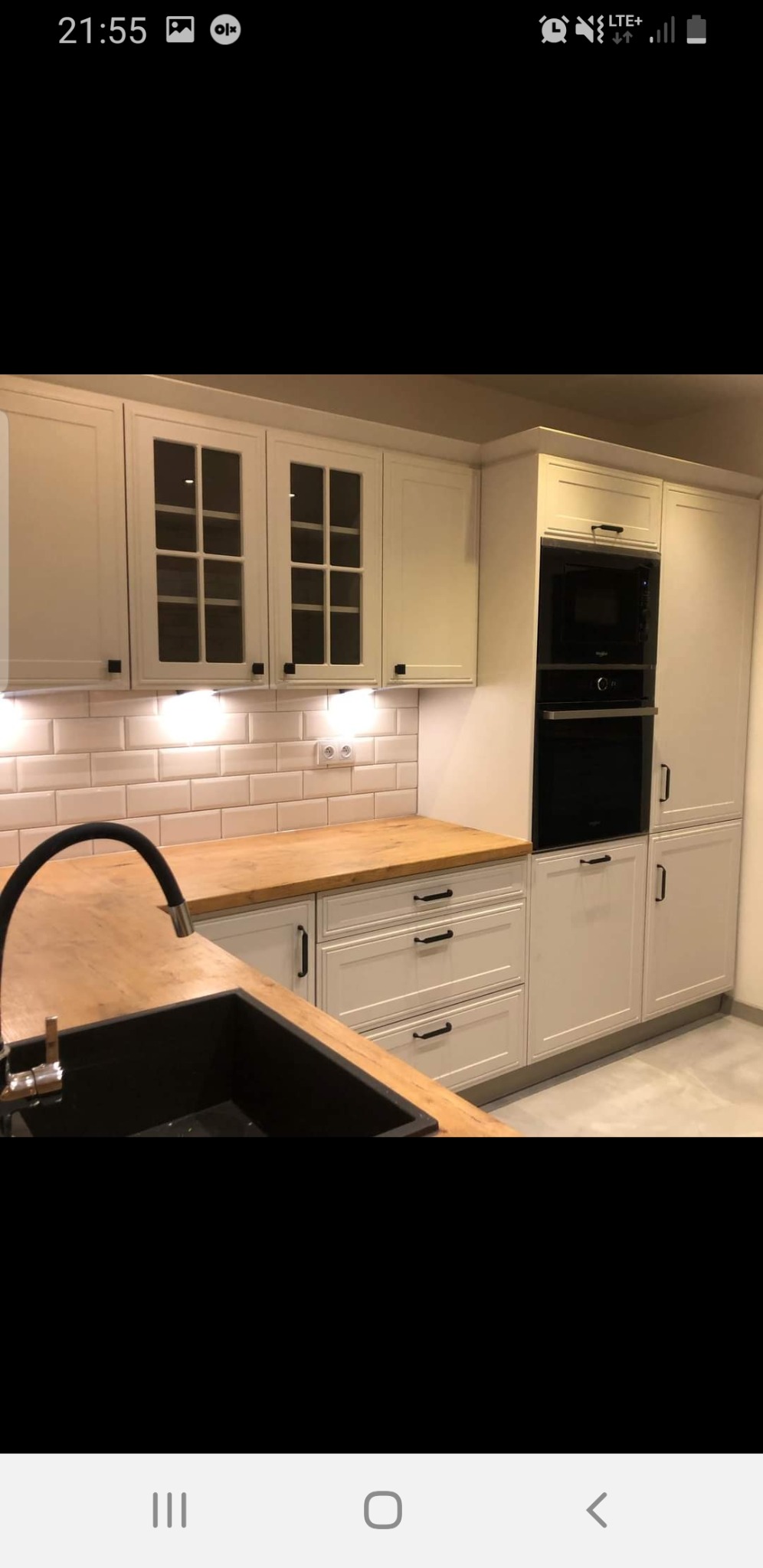 Nowoczesna kuchnia w odcieniach bieli z drewnianym blatem, czarnym zlewem i armaturą, oraz zabudowanymi piekarnikiem i mikrofalówką.