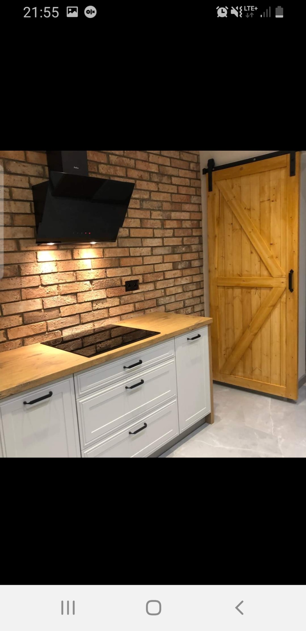 Wykończona kuchnia w Nowogrodźcu z ceglaną ścianą, białymi szafkami z drewnianym blatem, czarnym okapem i przesuwnymi drewnianymi drzwiami do spiżarni.