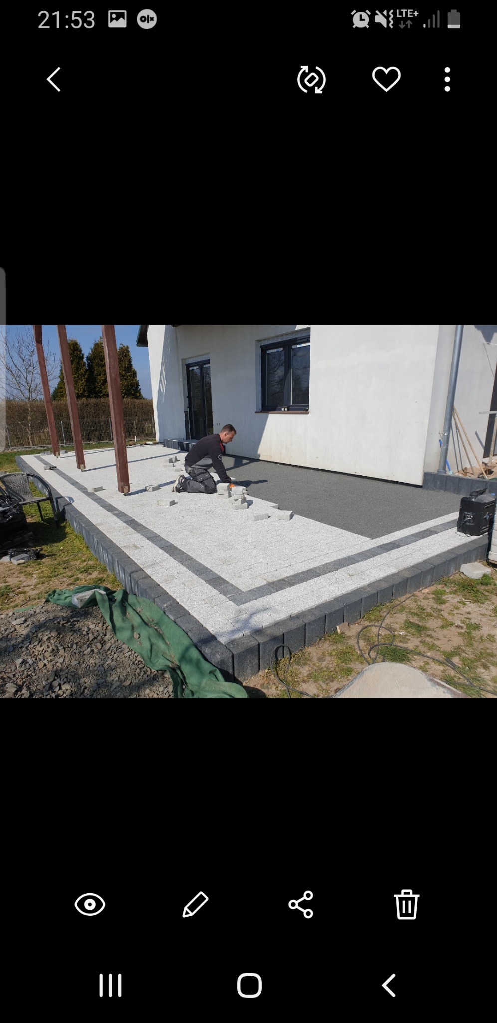 Pracownik układa jasnoszarą kostkę brukową na tarasie domu jednorodzinnego, obramowanym ciemniejszą kostką, częściowo na przygotowanym podłożu, z widocznymi elementami drewnianej konstrukcji pergoli.