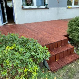 Brązowy drewniany taras z trzema schodkami, z roślinnością po bokach, widok na dom z otwartymi drzwiami balkonowymi i narzędzia na parapecie okna.