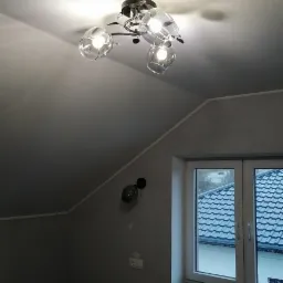 Nowoczesna lampa sufitowa z trzema szklanymi kloszami w szarym pokoju na poddaszu, widok na okno z widokiem na dachy.