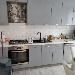 Jasnoszara kuchnia z białymi płytkami metra, czarną armaturą i piekarnikiem Beko; widoczna dekoracja ścienna z motywem uliczki.