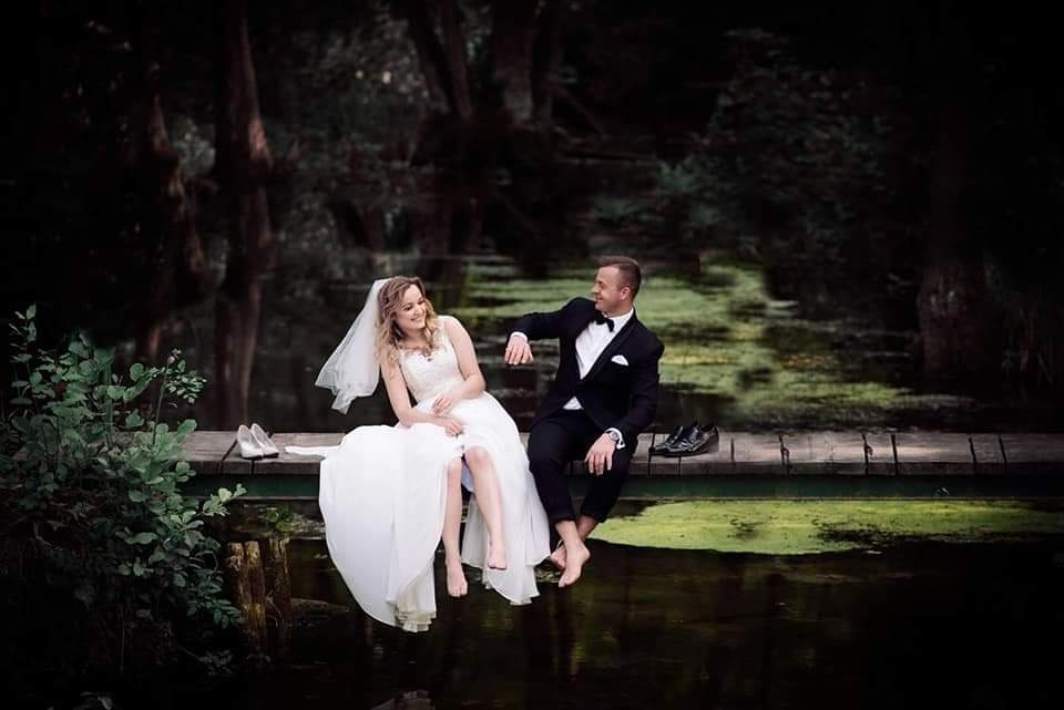 Para młoda siedzi boso na drewnianym pomoście nad wodą w otoczeniu drzew; panna młoda w białej sukni i welonie, pan młody w czarnym smokingu z muszką.