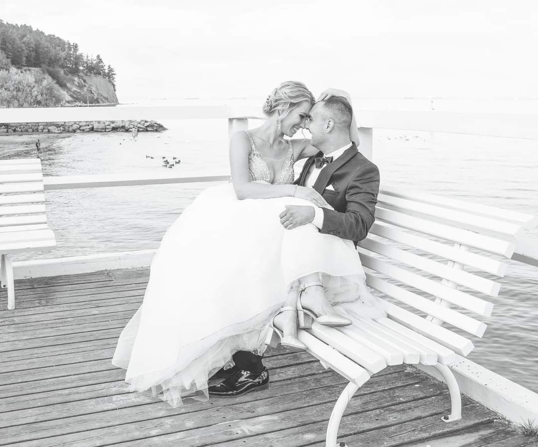 Czarno-białe zdjęcie uśmiechniętej pary młodej siedzącej na białej ławce na drewnianym molo z widokiem na morze i klif porośnięty drzewami w tle.