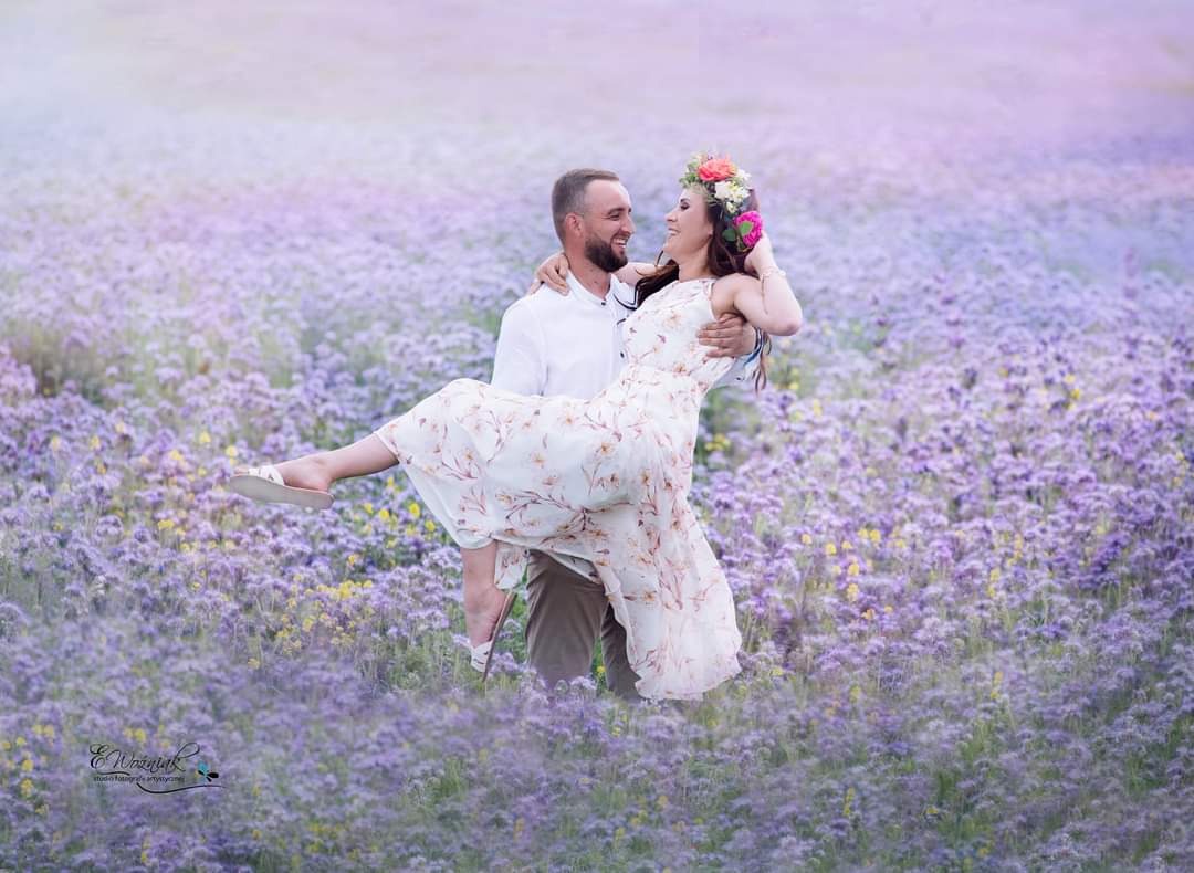 Uśmiechnięta para w objęciach na tle pola kwitnących, fioletowych kwiatów; kobieta w kwiecistej sukience i wianku na głowie, mężczyzna w białej koszuli.