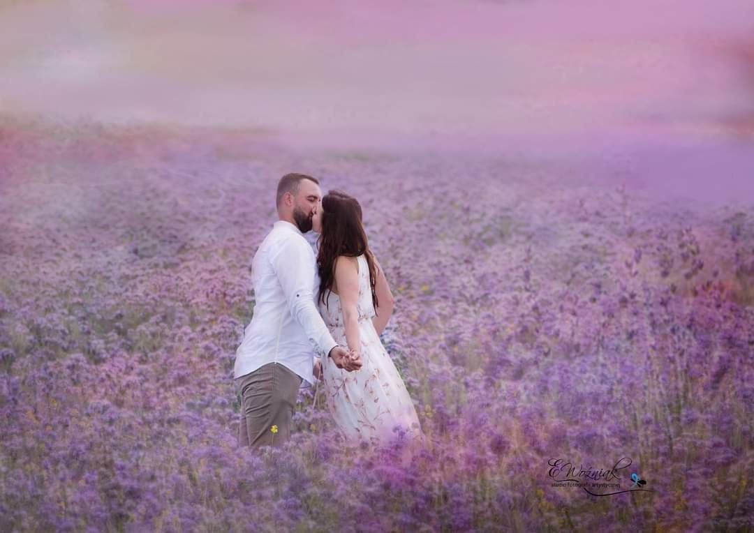 Zakochana para całuje się, stojąc pośród pola fioletowych kwiatów; mężczyzna w białej koszuli i beżowych spodniach, kobieta w kwiecistej sukience.