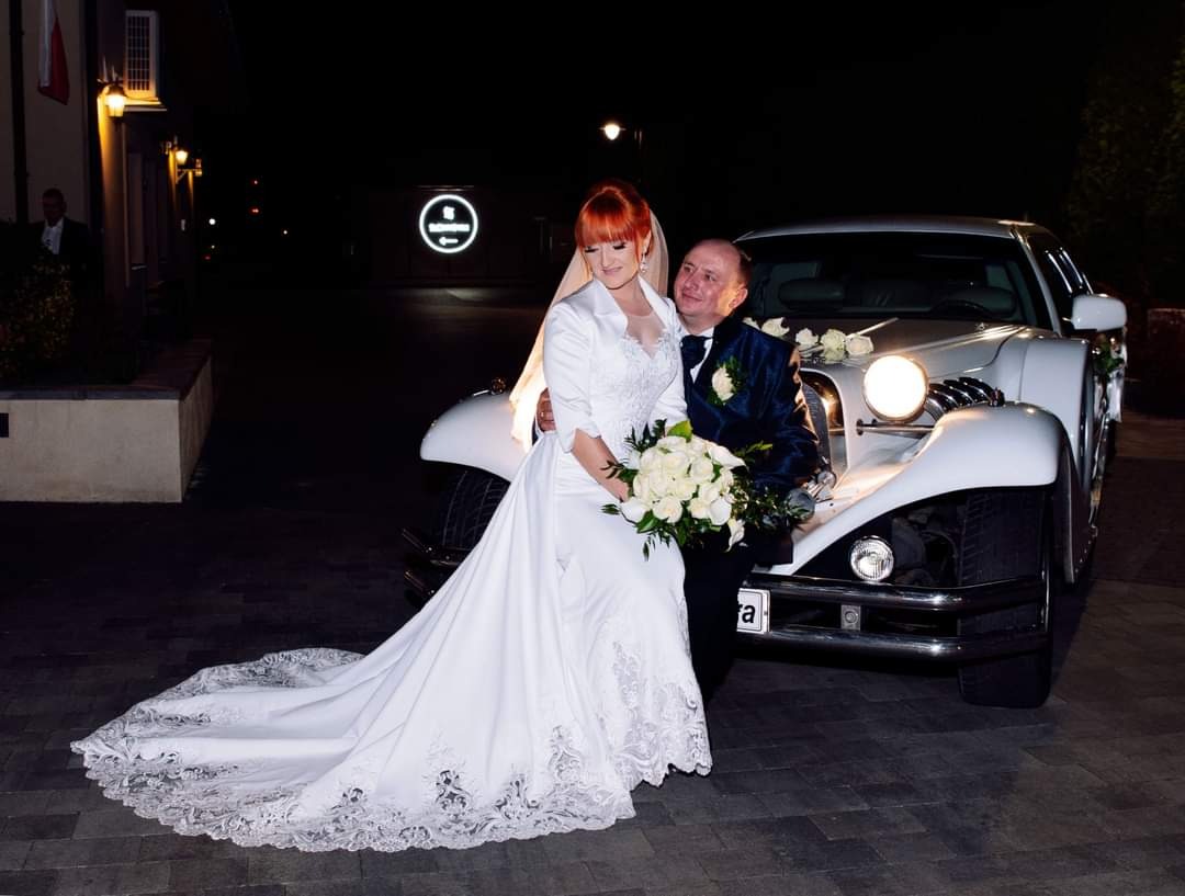 Para młoda pozuje przy białej limuzynie w nocy, panna młoda trzyma bukiet białych róż, w tle budynek z oświetlonym logo.