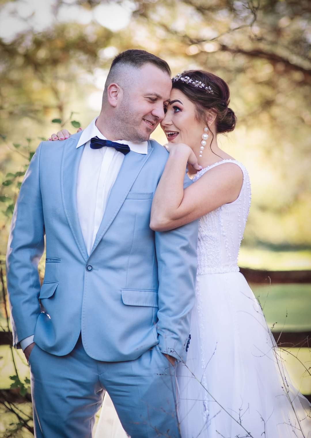 Uśmiechnięta para młoda w pastelowych strojach: on w błękitnym garniturze z muchą, ona w białej sukni z perłowymi kolczykami i tiarą, opierająca się o jego ramię na tle zieleni.