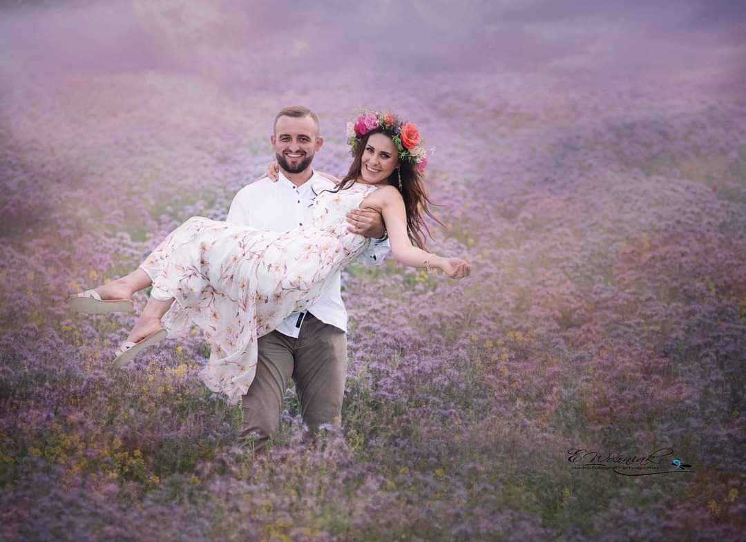 Uśmiechnięty mężczyzna trzyma na rękach kobietę w kwiecistej sukience i wianku na głowie, stoją oboje na fioletowym polu kwiatów, delikatne światło, romantyczna sesja.