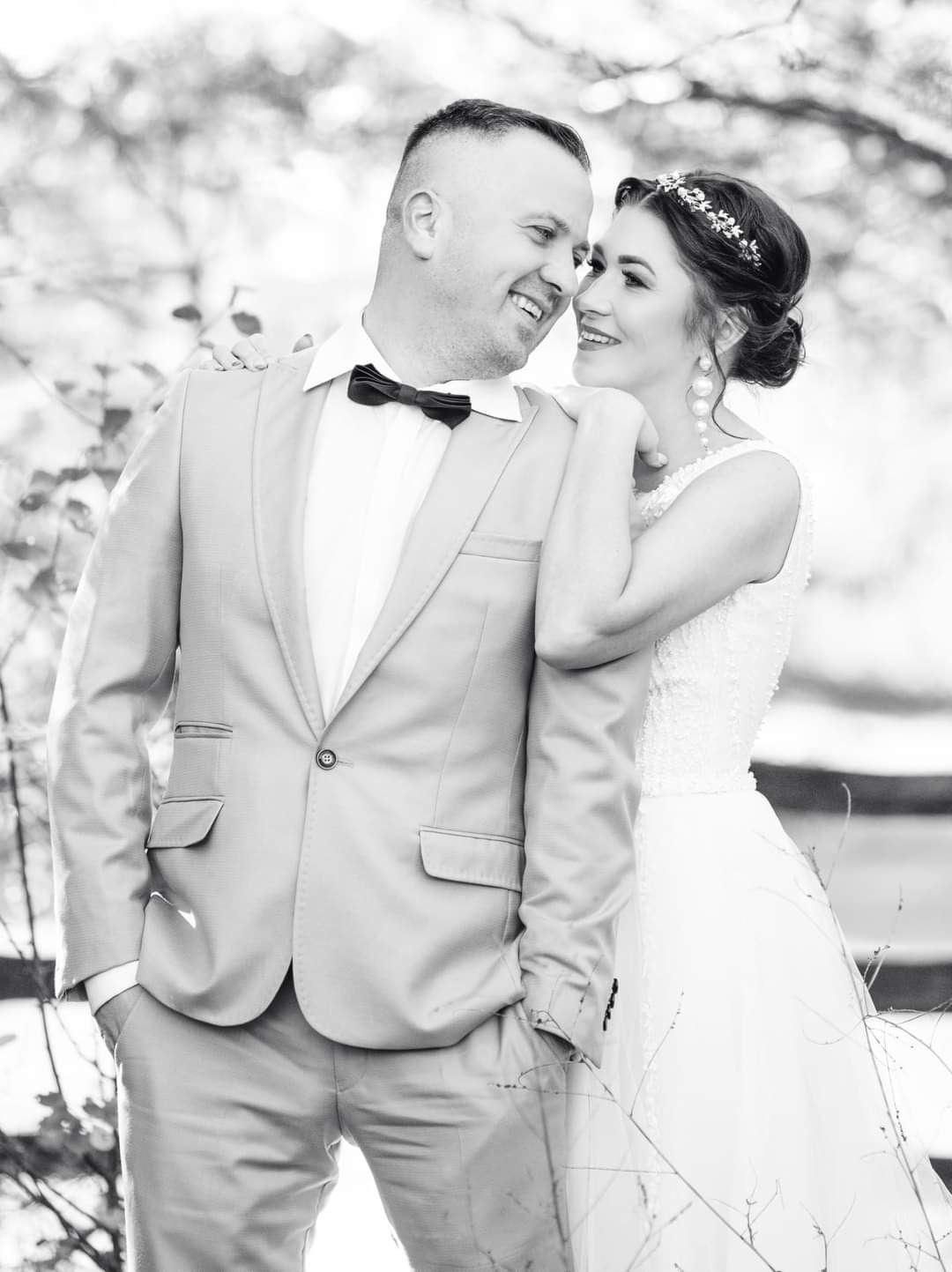 Czarno-białe zdjęcie uśmiechniętej pary młodej: panna młoda opiera głowę na ramieniu pana młodego w jasnym garniturze z muchą, w tle rozmyte drzewa i woda.