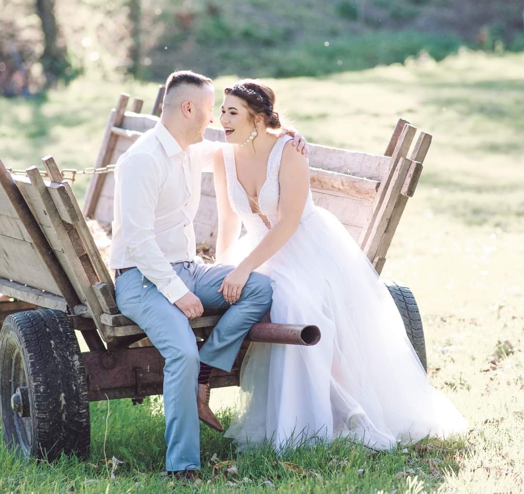 Uśmiechnięta para młoda siedzi na starym drewnianym wozie na tle zielonej łąki w słoneczny dzień. Panna młoda w białej sukni z głębokim dekoltem i tiarą, pan młody w białej koszuli i błękitnych...