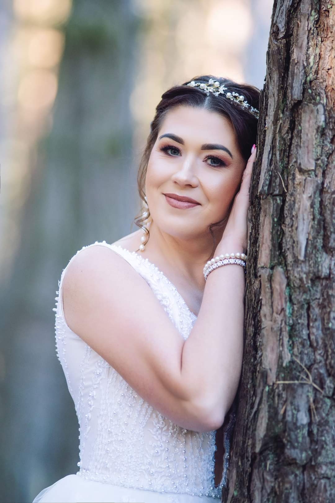 Uśmiechnięta panna młoda w białej sukni z perłami i tiarą, opierająca się o korę drzewa w leśnej scenerii.