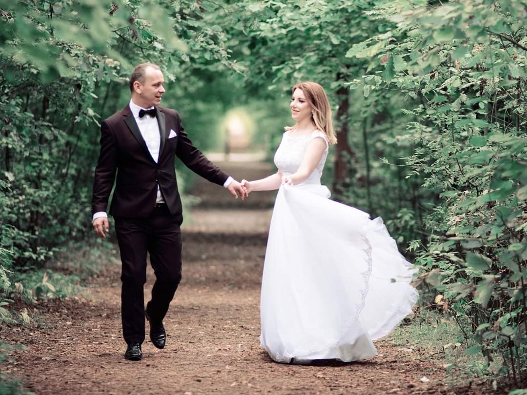 Para młoda, on w ciemnym garniturze z muchą, ona w białej sukni, idą trzymając się za ręce leśną ścieżką w kierunku światła.