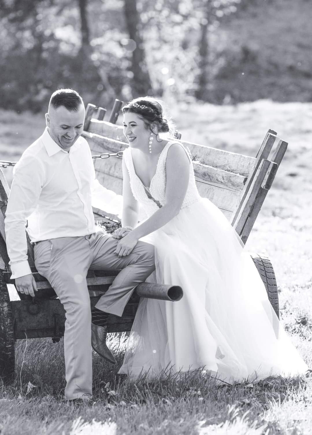 Czarno-białe zdjęcie uśmiechniętej panny młodej w koronkowej sukni i pana młodego siedzących na starym, drewnianym wozie na tle łąki i drzew.