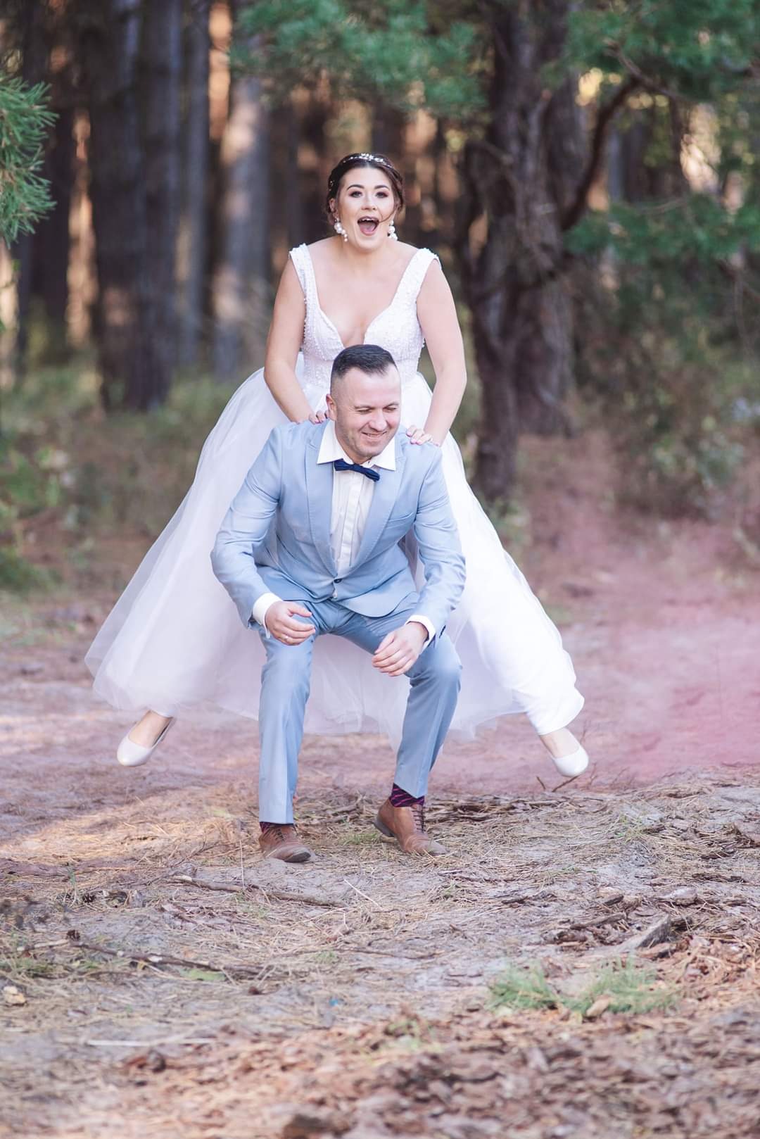 Panna młoda w białej sukni, z szeroko otwartymi ustami, wskakuje na barana panu młodemu w błękitnym garniturze, który kuca na leśnej ścieżce.