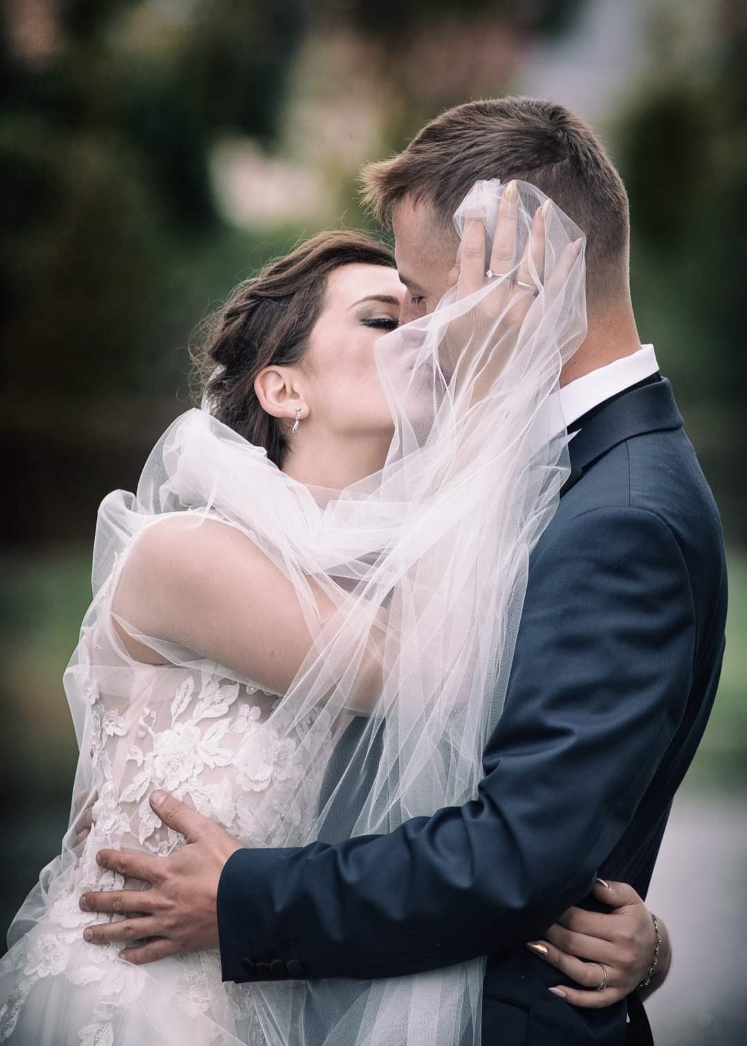 Pocałunek nowożeńców: panna młoda w koronkowej sukni i welonie, pan młody w granatowym garniturze, widoczny pierścionek zaręczynowy na palcu dłoni trzymającej welon.
