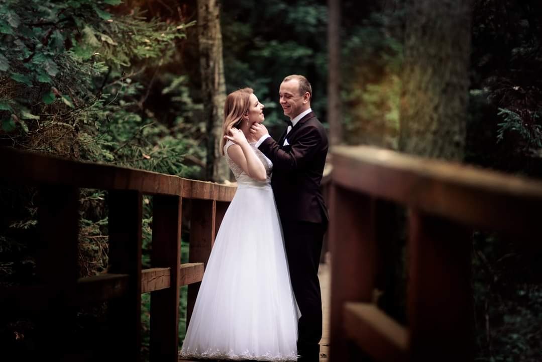 Para młoda, on w czarnym garniturze z muszką, ona w białej sukni ślubnej, stoi na drewnianym mostku w lesie, patrząc na siebie z uśmiechem.