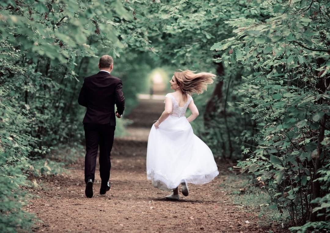 Para młoda biegnie ścieżką w lesie; panna młoda w zwiewnej sukni z odkrytymi plecami, jej włosy unoszą się w ruchu, a pan młody w ciemnym garniturze, oboje oddalają się od kamery.
